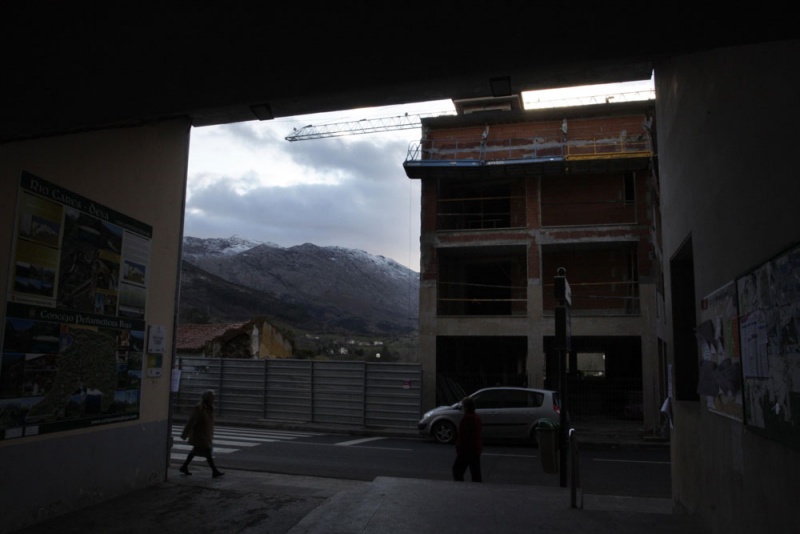 Dos mujeres se cruzan delante del Ayuntamiento de Panes, con un edificio en construcción, detrás, y las montañas nevadas, al fondo