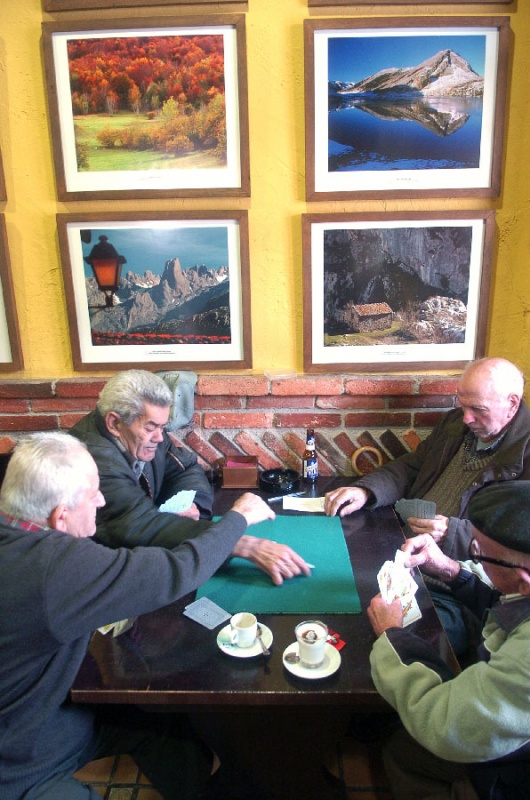 De izquierda a derecha, Ángel Alonso, José Díaz, José Sarrapio y Salvador Sánchez juegan al tute en el café Cares, ante una exposición de fotos de los Picos hechas por Alfredo Fernández