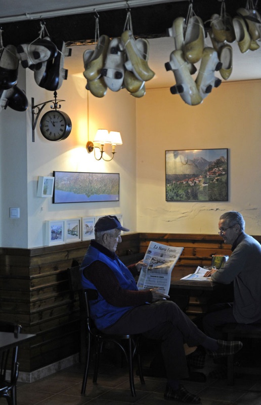 Félix Fernández lee LA NUEVA ESPAÑA junto a José Alonso Simón en el bar-tienda Casa Chili, tras el muestrario de madreñas y ante una fotografía de Sames