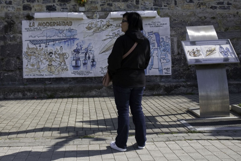 Una mujer observa uno de los paneles sobre la historia de Ribadesella dibujados por Antonio Mingote en el paseo de la Grúa.